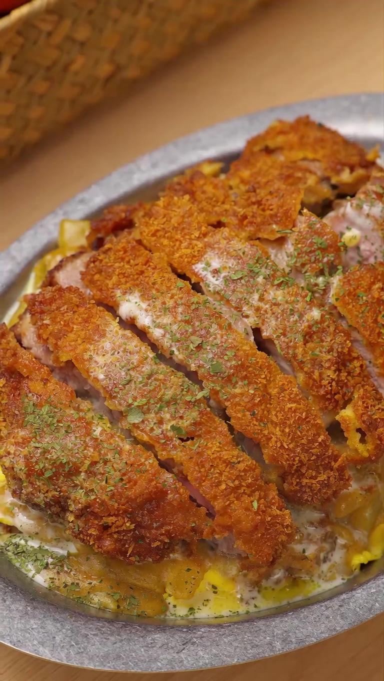 Chrupiący, pokrojony w plastry kotlet schabowy ułożony na miękkiej jajecznicy i ryżu, polany bogatym kokosowym curry i kremowym sosem serowym.