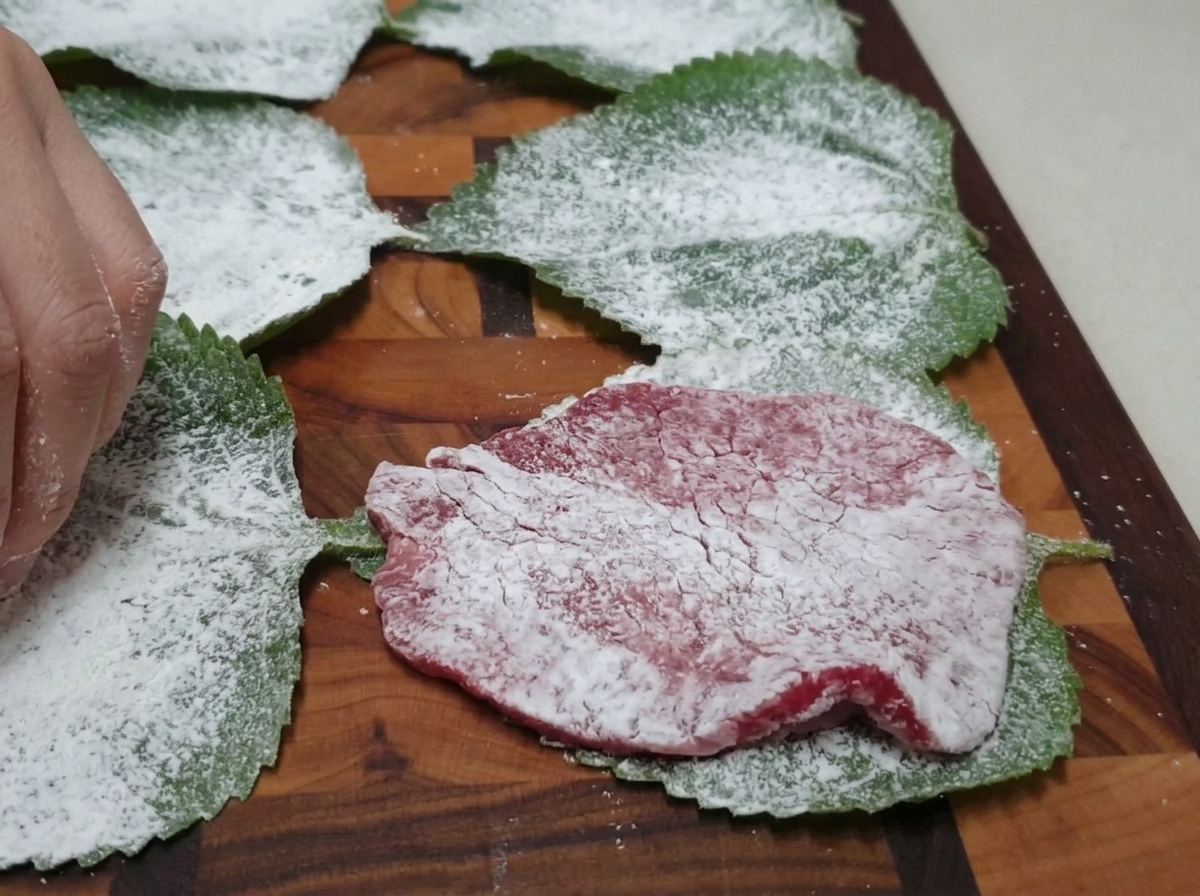 Oprószony mąką plaster wołowiny układany na oprószonym liściu pachnotki na desce do krojenia.