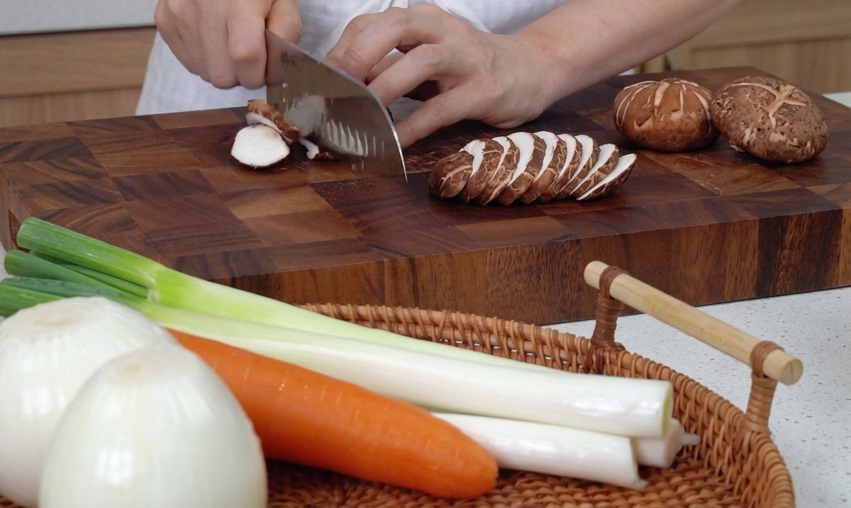 Dłonie krojące świeże grzyby shiitake nożem kuchennym na drewnianej desce.