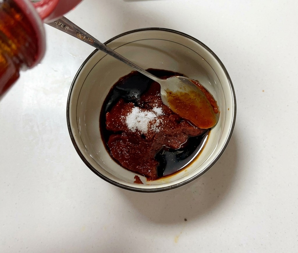 Mała miseczka wypełniona pastą gochujang, cukrem i sosem sojowym, mieszana metalową łyżką.