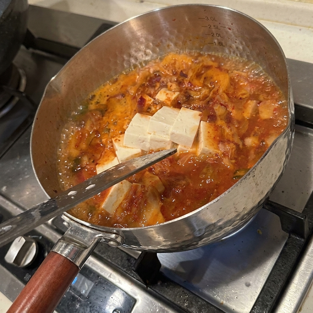 Różnorodne świeże składniki, w tym tofu, grzyby enoki i kimchi gotujące się w pikantnym, czerwonym wywarze.