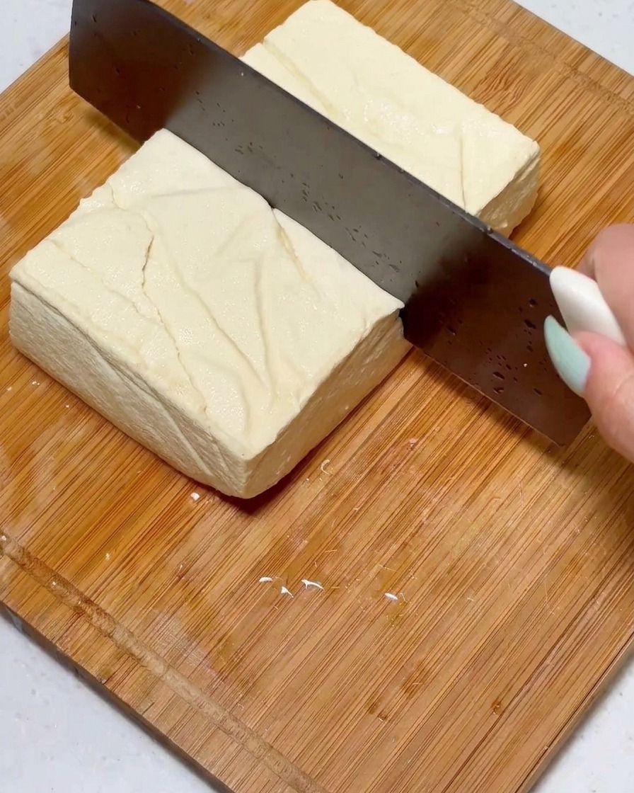 Krojenie kwadratowego bloku jędrnego tofu na równe kawałki za pomocą dużego noża na drewnianej powierzchni.