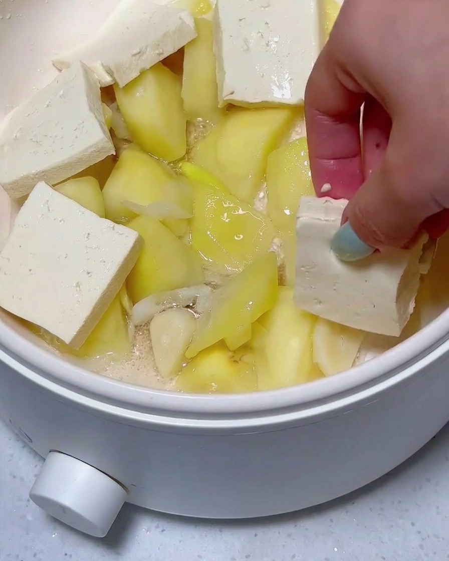 Warstwy plastrów tofu i mielonki ułożone na kawałkach ziemniaków na patelni.