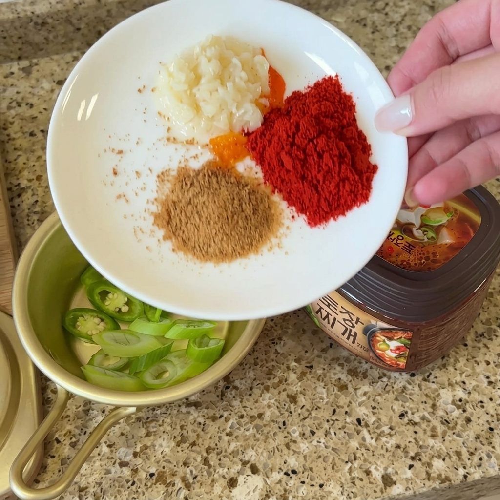 Biały talerz z małymi porcjami posiekanego czosnku, chili w proszku, bulionu wołowego i sproszkowanej pachnotki obok pojemnika z pastą doenjang.
