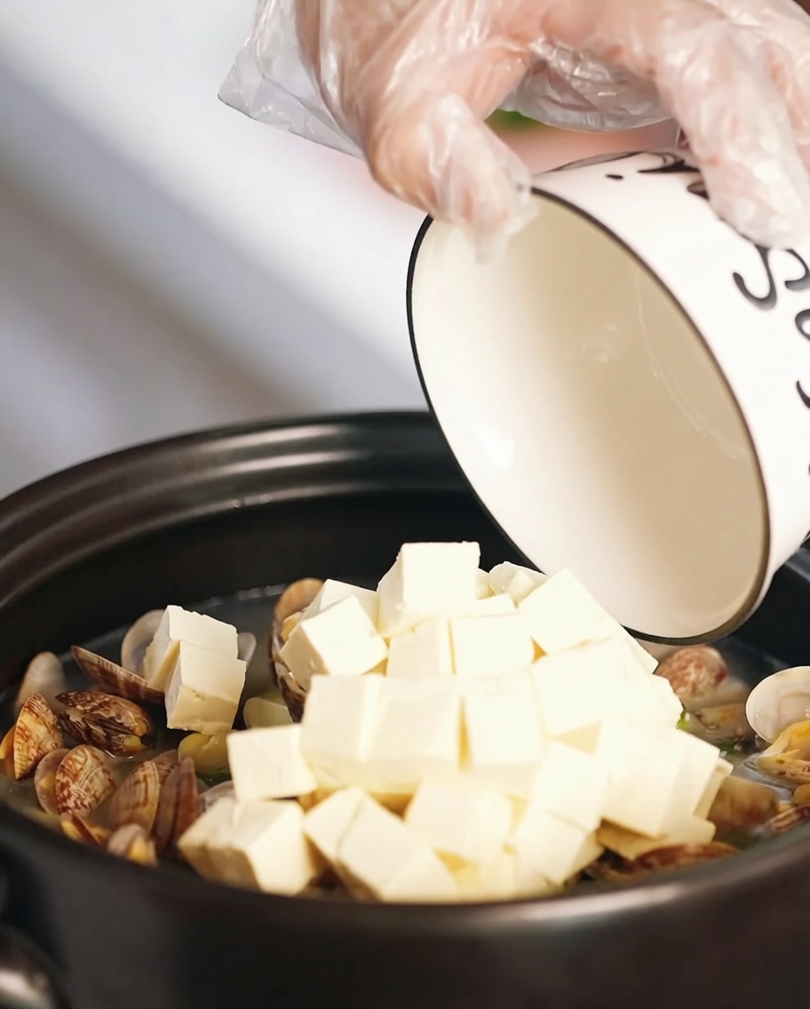 Kostki miękkiego tofu i kawałki smażonego jajka dodawane do zupy z małży w garnku.