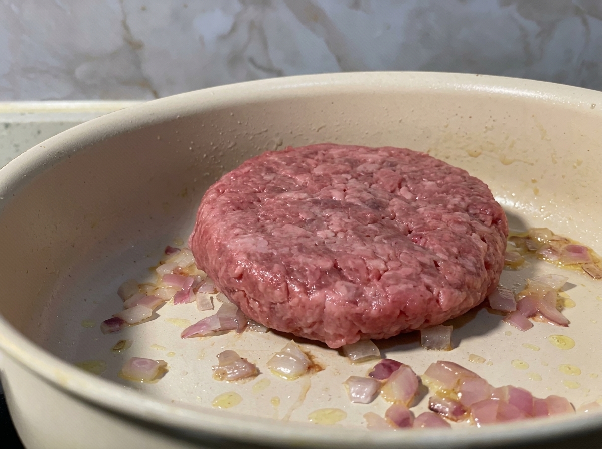 Surowy kotlet z mielonej wołowiny położony na patelni obok podsmażonej czerwonej cebuli.