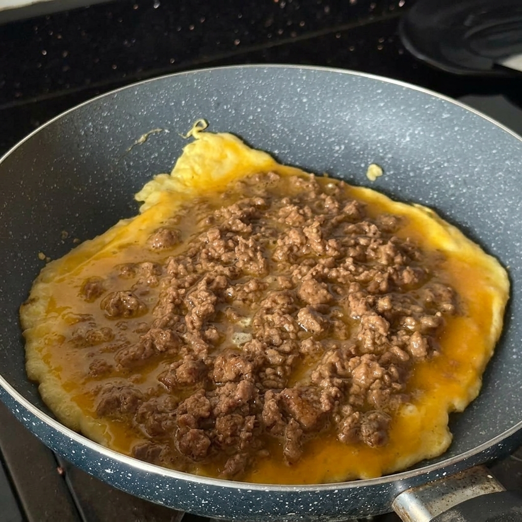 Omlet jajeczny smażący się na patelni, obficie posypany wołowiną.