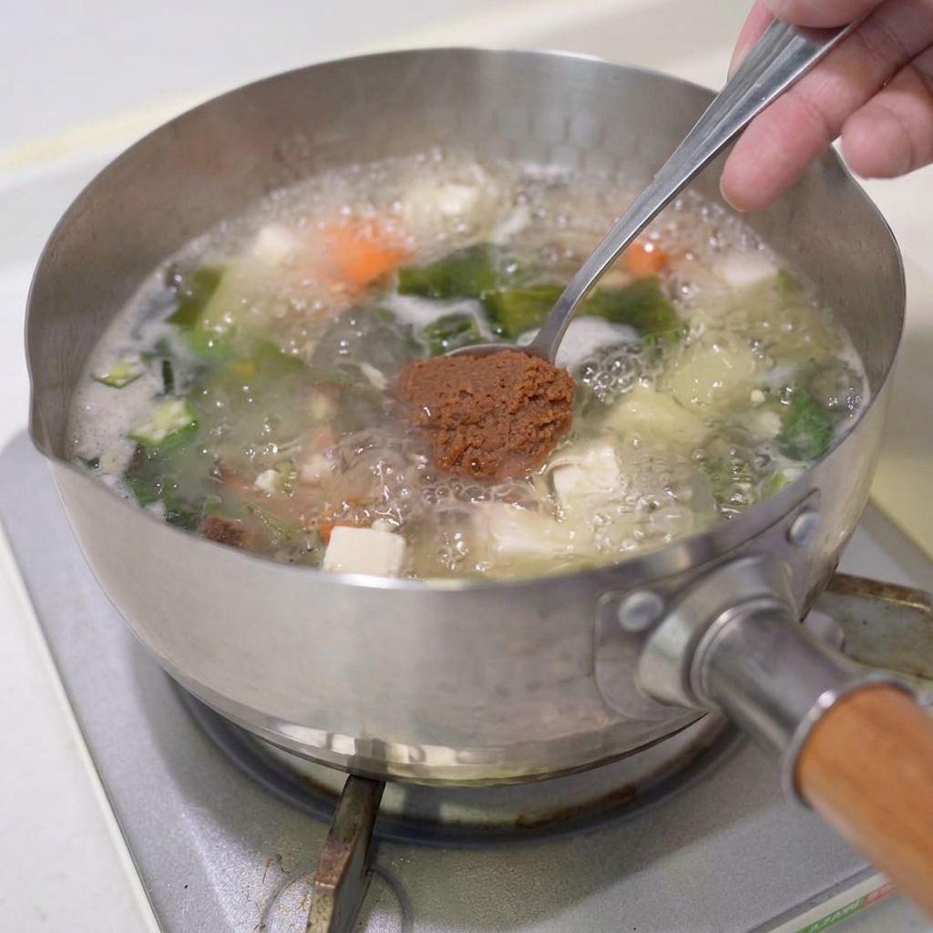Łyżka brązowej pasty miso zanurzana w gotującej się zupie.
