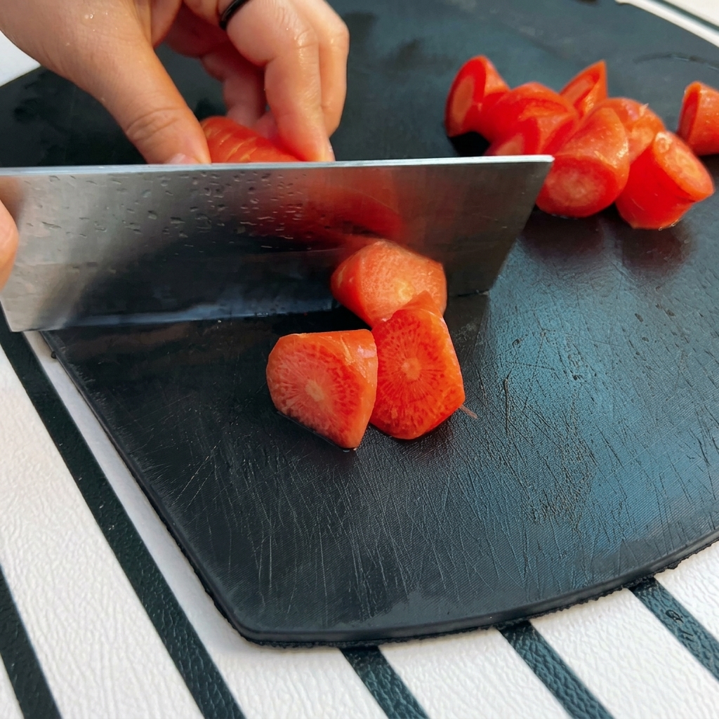 Ręka trzymająca tasak krojąca pomarańczową marchew na kawałki metodą roll-cut na czarnej desce.