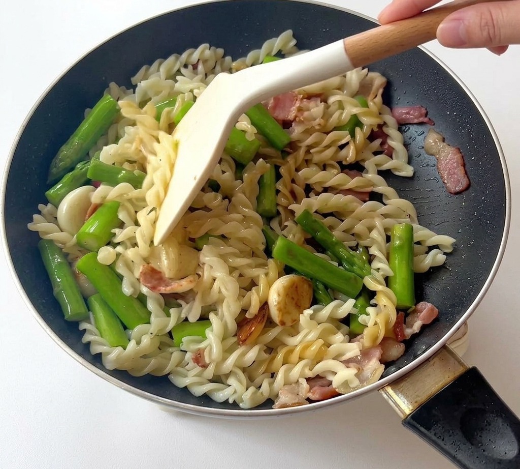 Spatuła mieszająca makaron fusilli ze szparagami i boczkiem na patelni.