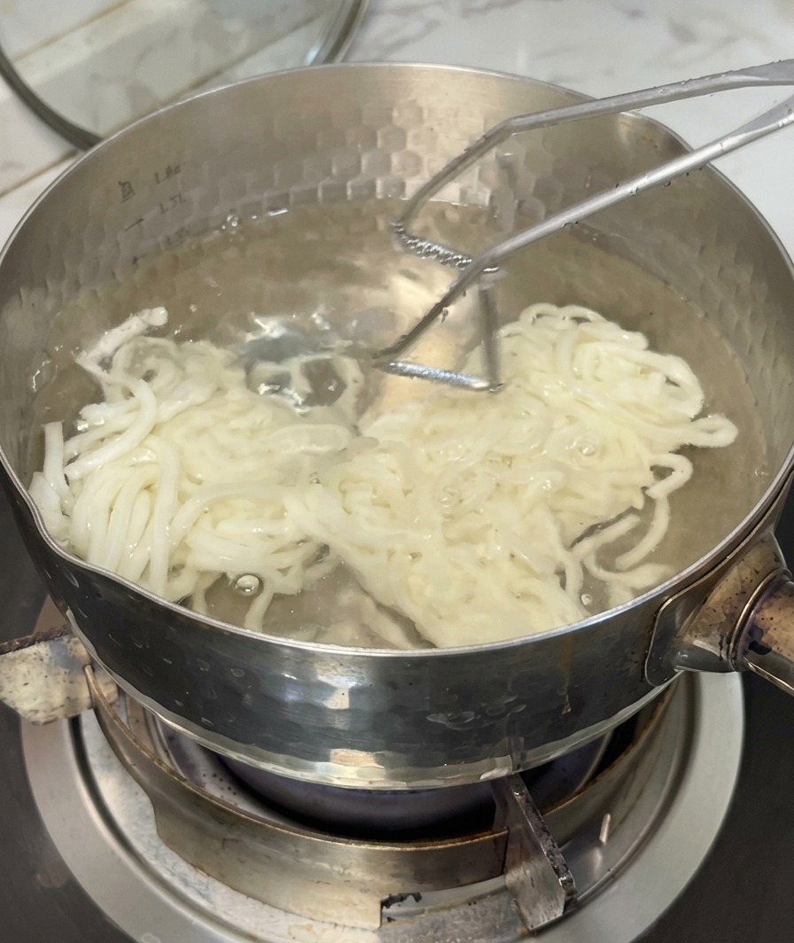 Sprężysty makaron udon wyjmowany z garnka z wrzącą wodą za pomocą sitka.