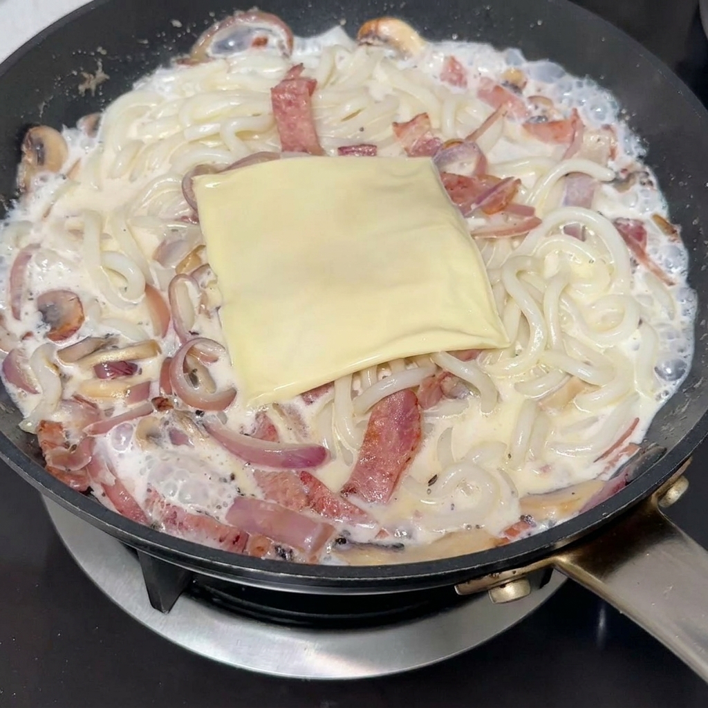 Plasterek sera topionego leżący na wierzchu kremowego makaronu udon.