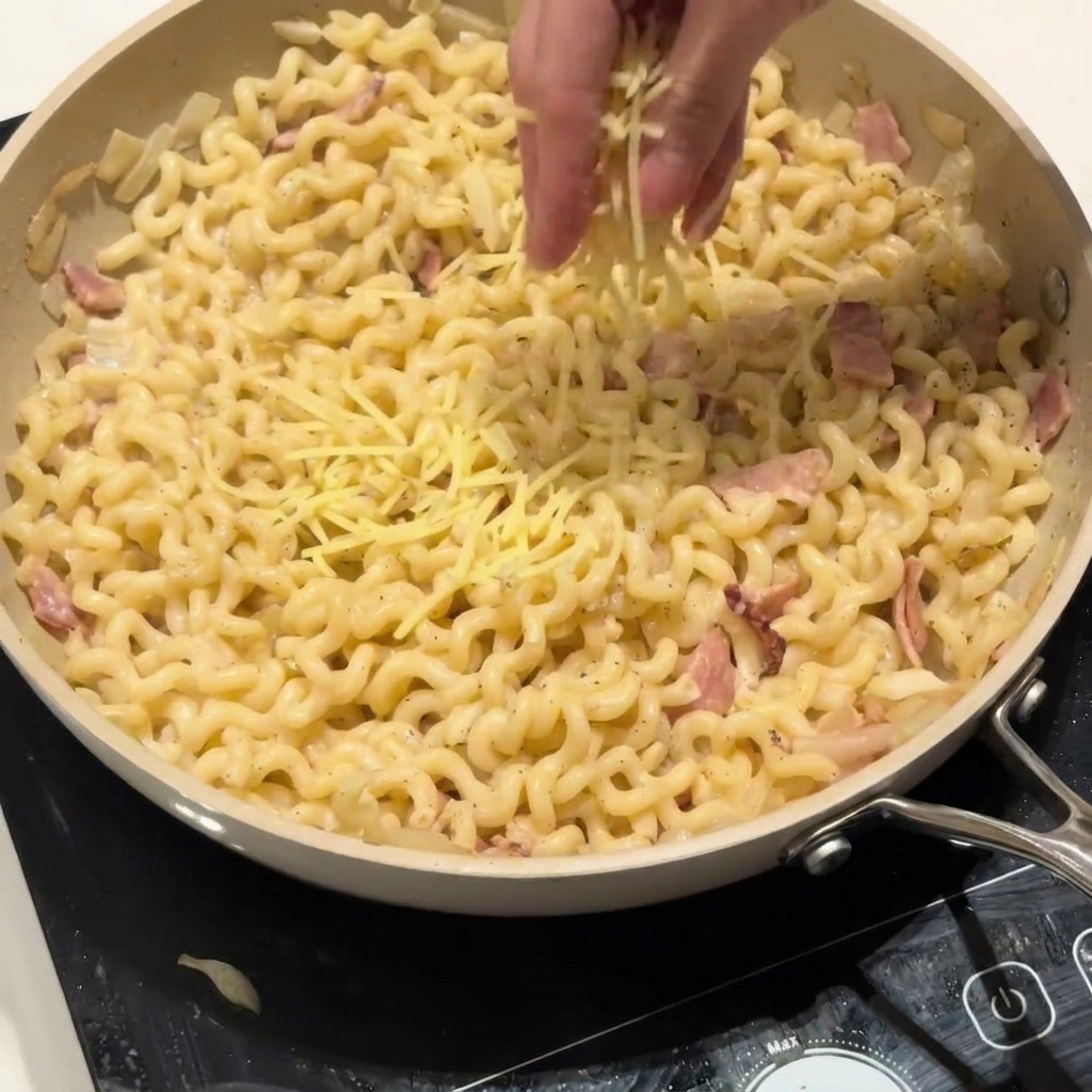 Ręka posypująca starty parmezan na patelnię z kremowym makaronem.