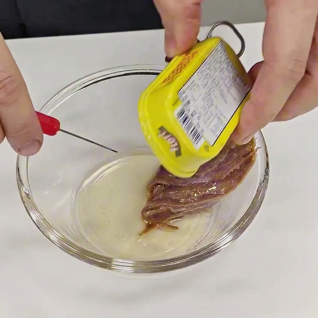 Dłonie ostrożnie wyjmujące filet anchois z żółtej puszki i wkładające go do małej, przezroczystej miseczki.