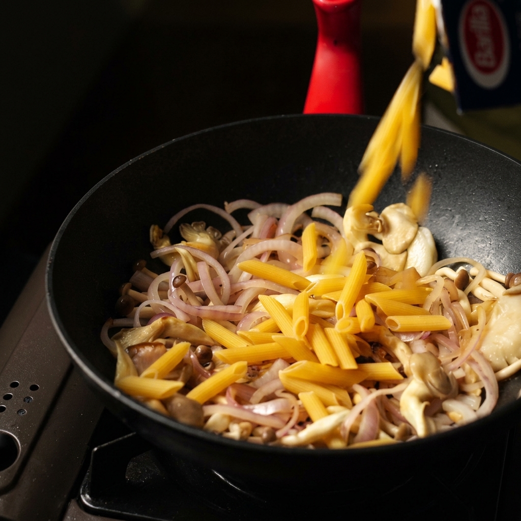Suchy makaron penne wsypywany do patelni z podsmażonymi grzybami i cebulą.