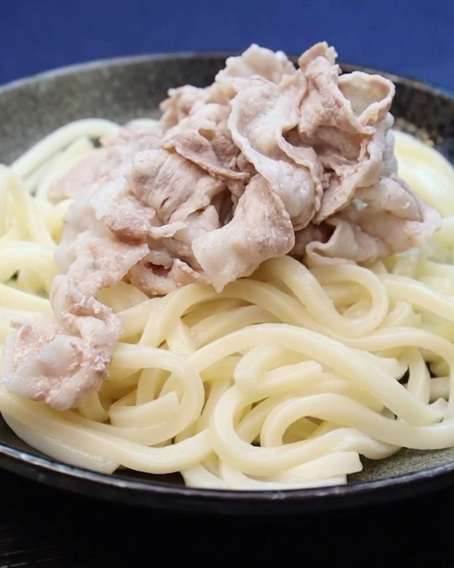 Ugotowane plastry boczku układane na porcji makaronu udon.