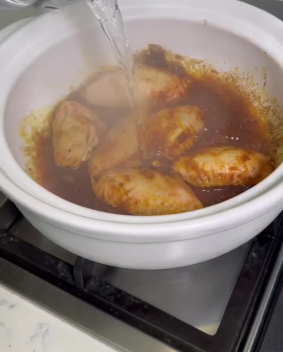 Czysta woda wlewana z naczynia do bulgoczącego, glinianego garnka wypełnionego skrzydełkami z kurczaka w curry.
