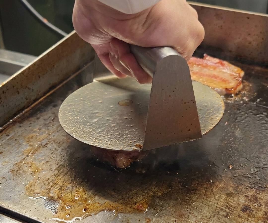 Metalowa praska do burgerów spłaszczająca kulkę wołowiny Wagyu na gorącej powierzchni.