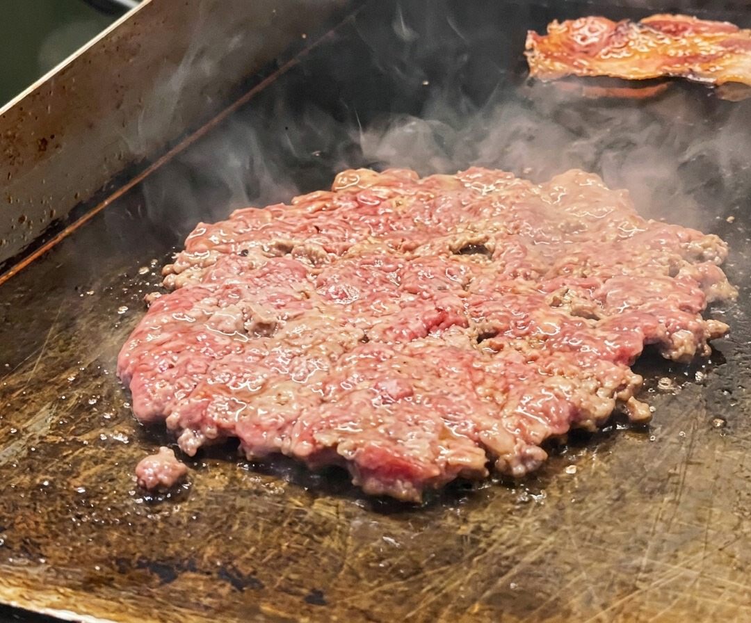 Cienki kotlet wołowy smażący się na patelni, posypany solą i przykryty plastrami cebuli.