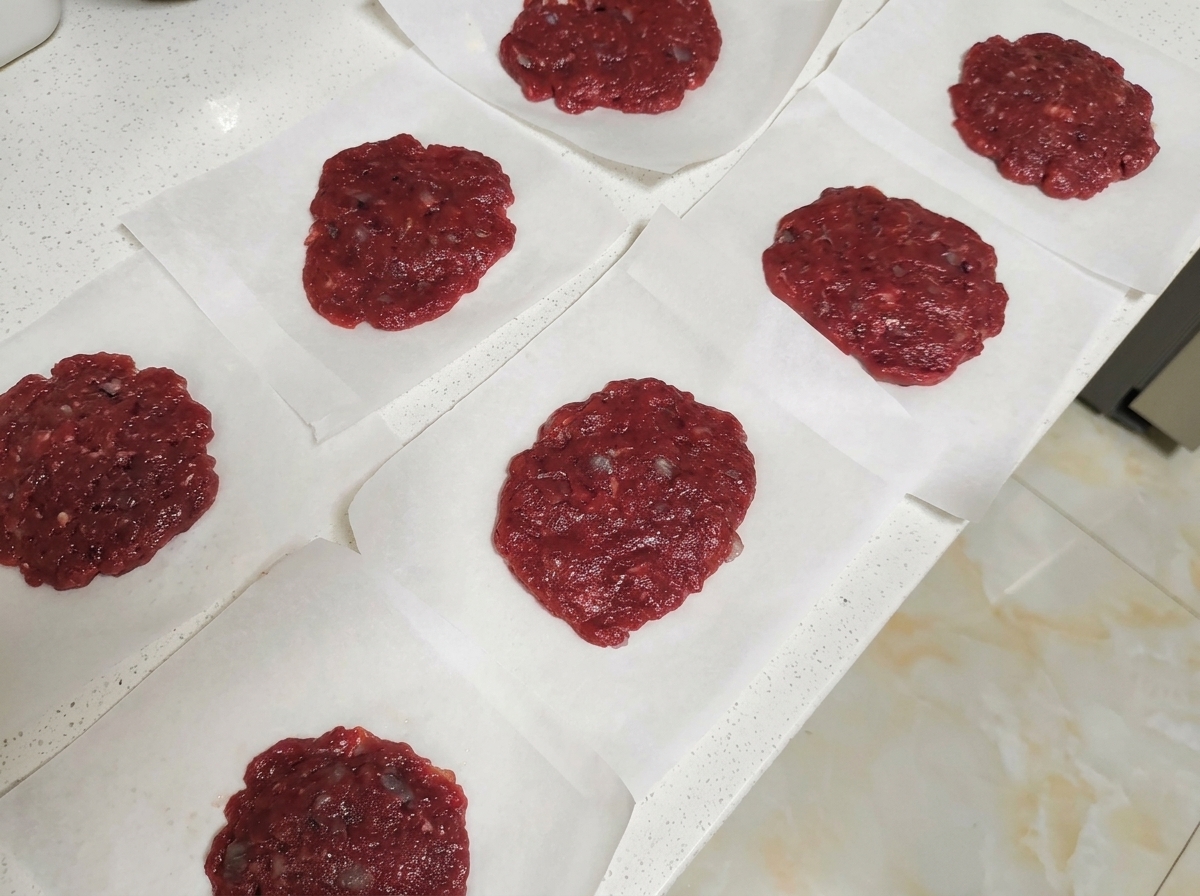 Kilka surowych okrągłych kotletów wołowych umieszczonych na kwadratach papieru do pieczenia na blacie.