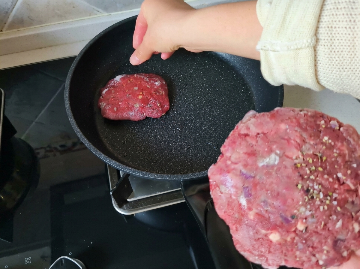 Ręka trzymająca doprawiony, surowy kotlet wołowy i wkładająca go na rozgrzaną, lekko natłuszczoną patelnię.