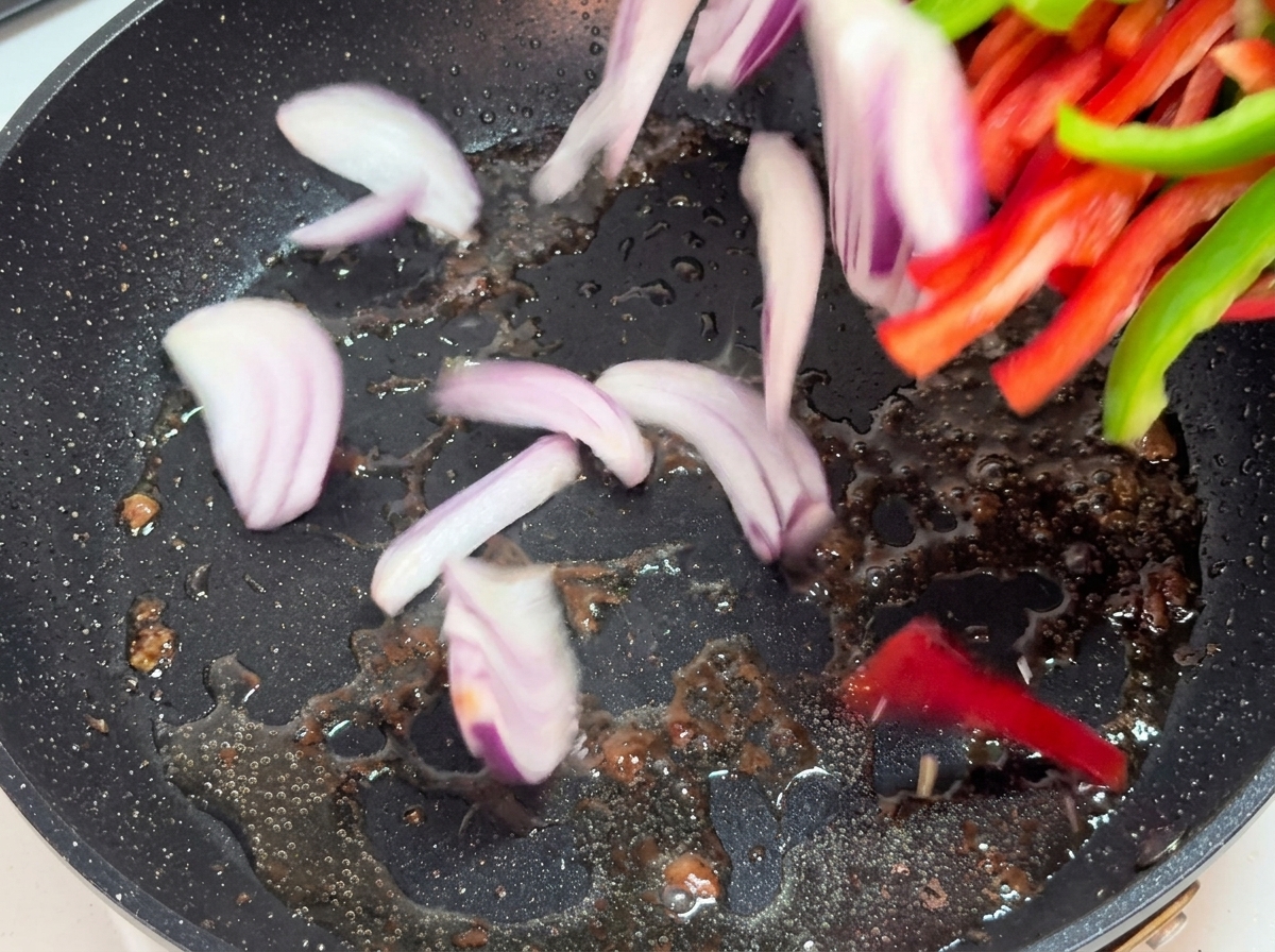 Paski cebuli i papryki smażące się na gorącej patelni.
