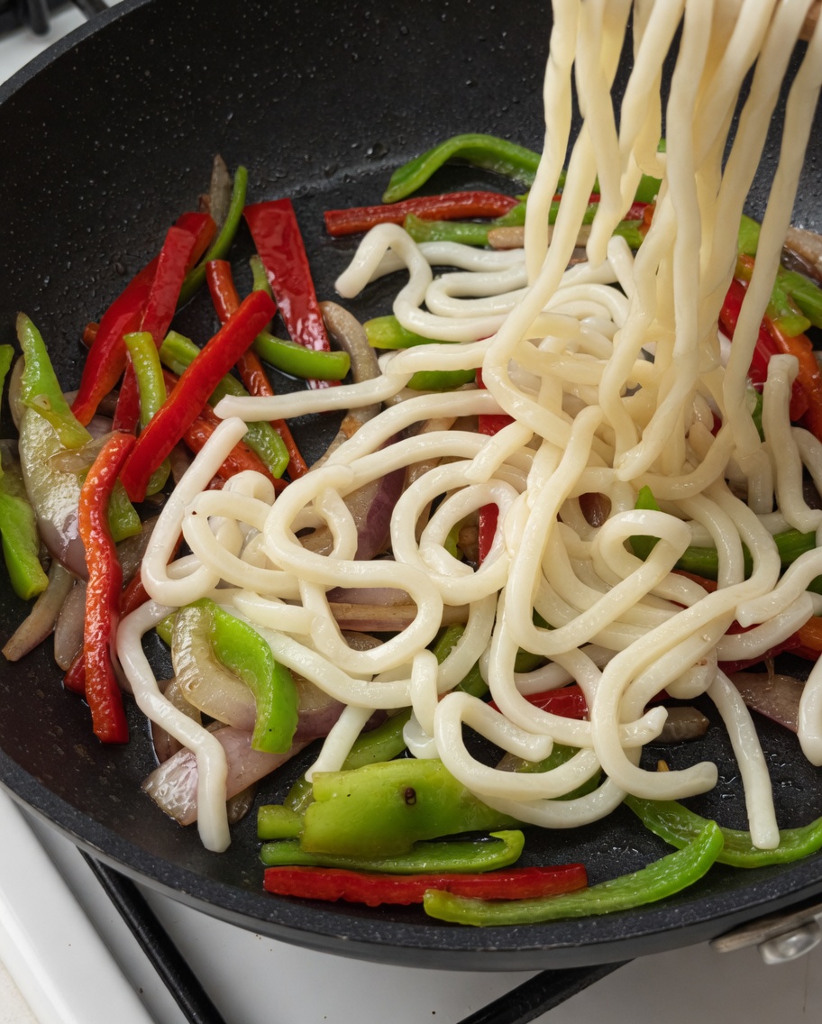 Makaron udon i paski wołowiny dodawane na patelnię z warzywami.