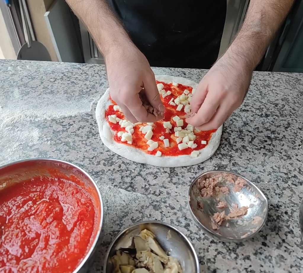 Dodawanie płatków tuńczyka i plasterków karczochów na pizzę jako finalnych dodatków.