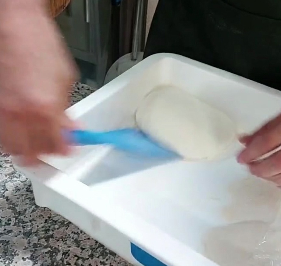 Porcja ciasta do pizzy wyjmowana z pojemnika za pomocą niebieskiej plastikowej skrobaczki.