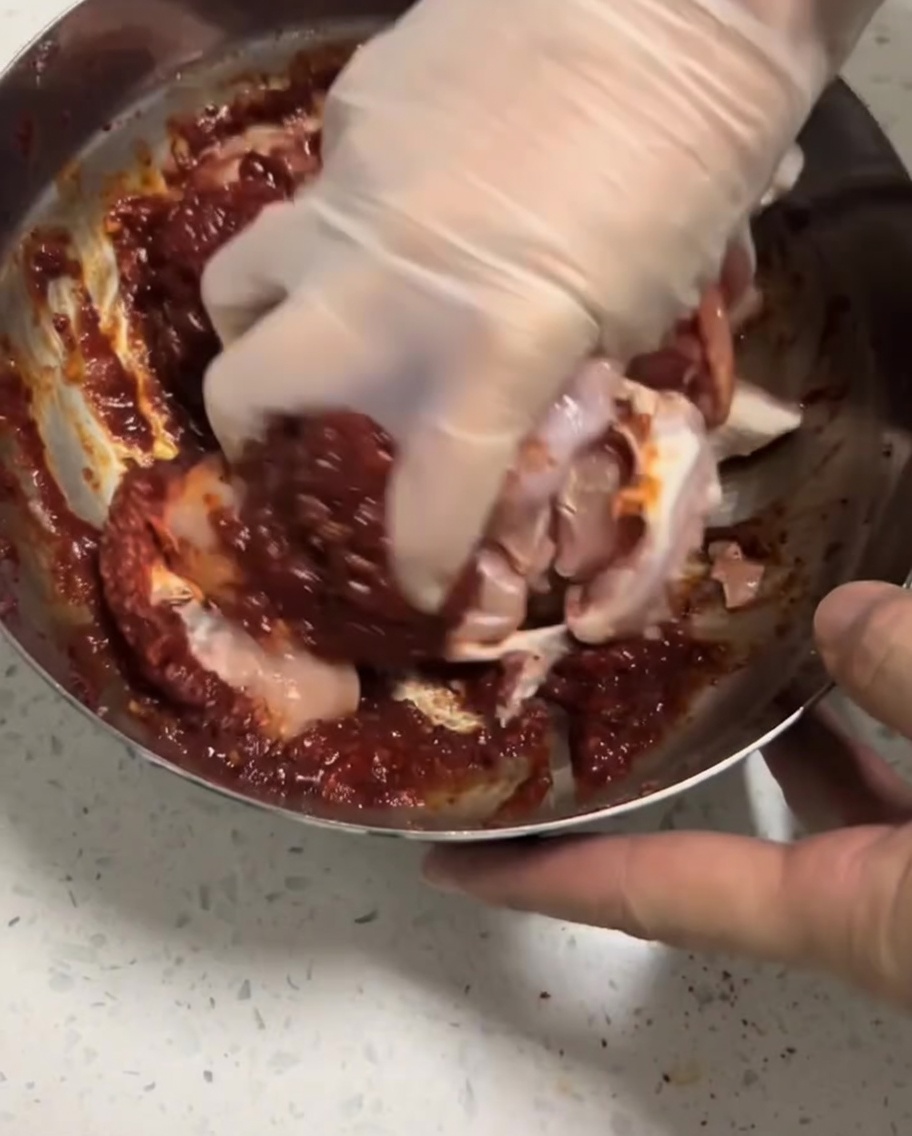 Surowe kawałki kurczaka nacierane czerwoną marynatą przez dłoń w rękawiczce kuchennej.