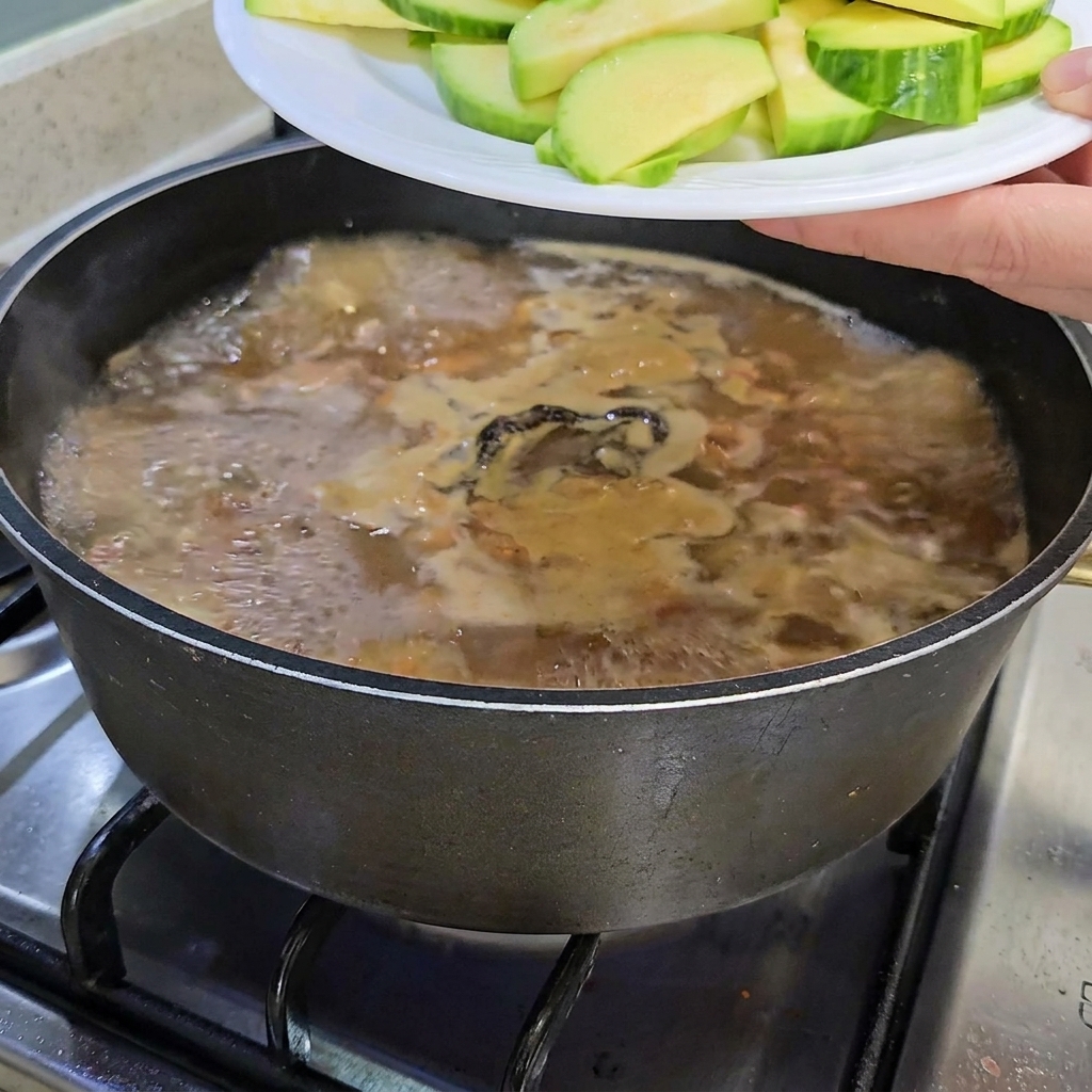 Pokrojona cukinia i pęczek grzybów enoki dodawane do garnka z gulaszem.