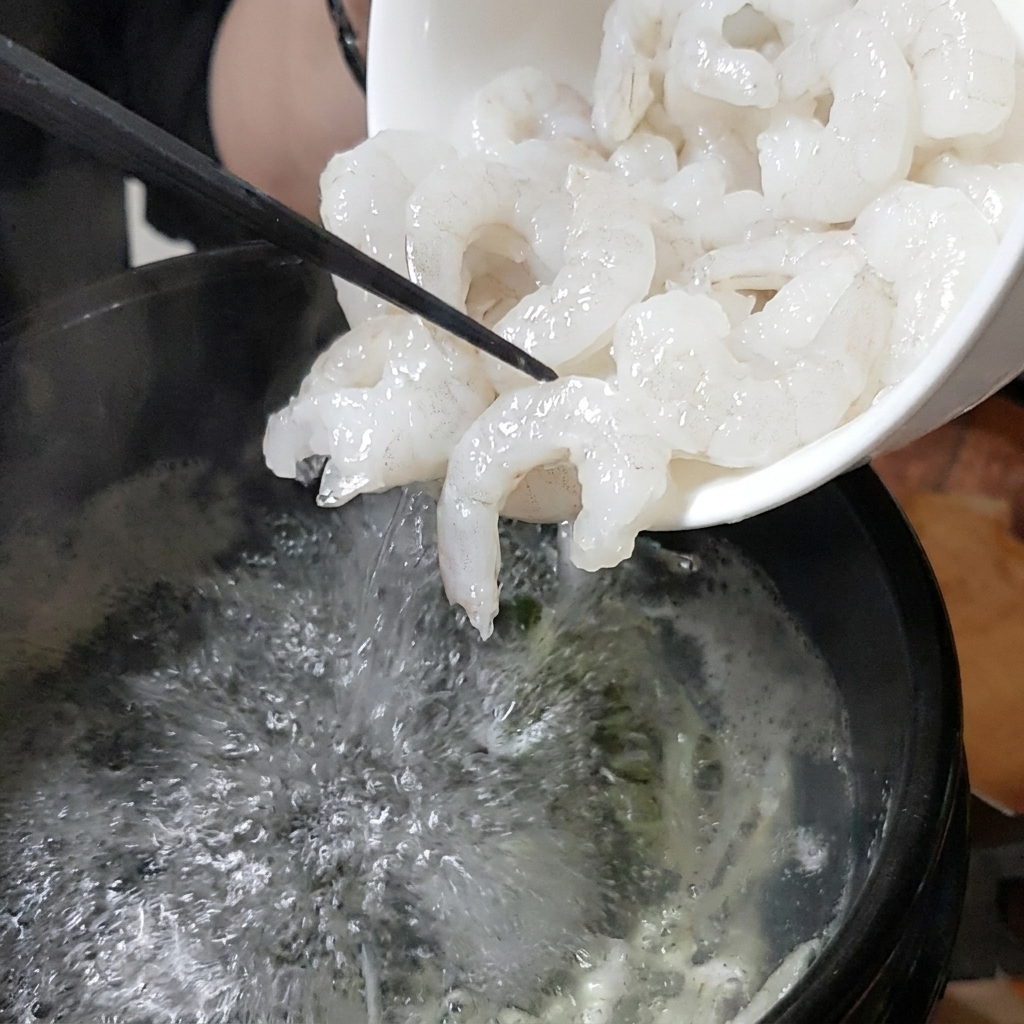 Biała miska z której świeże krewetki są wsypywane do buzującego kamiennego garnka z bulionem.