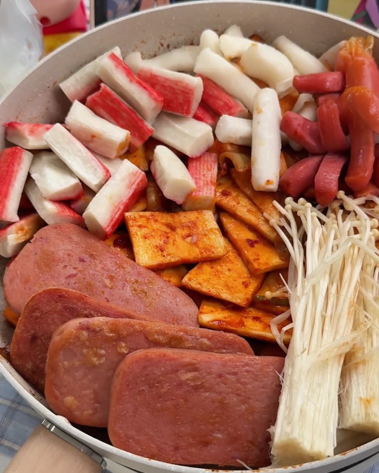 Patelnia z ładnie ułożonymi sekcjami mielonki, paluszków krabowych, kiełbasek, ciasteczek ryżowych, ciasteczek rybnych i grzybów enoki.
