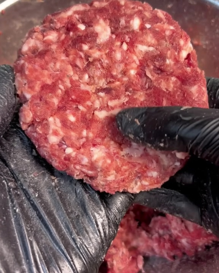 Dłoń w czarnej rękawiczce formująca doprawioną wołowinę w okrągły kotlet.