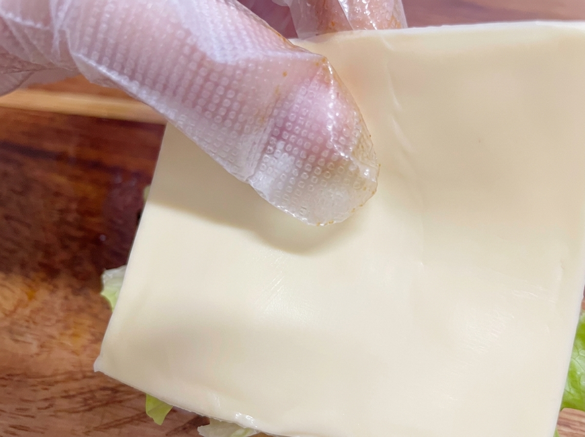 Dłoń w plastikowej rękawiczce ostrożnie kładąca kwadratowy plasterek białego sera na składanym burgerze.