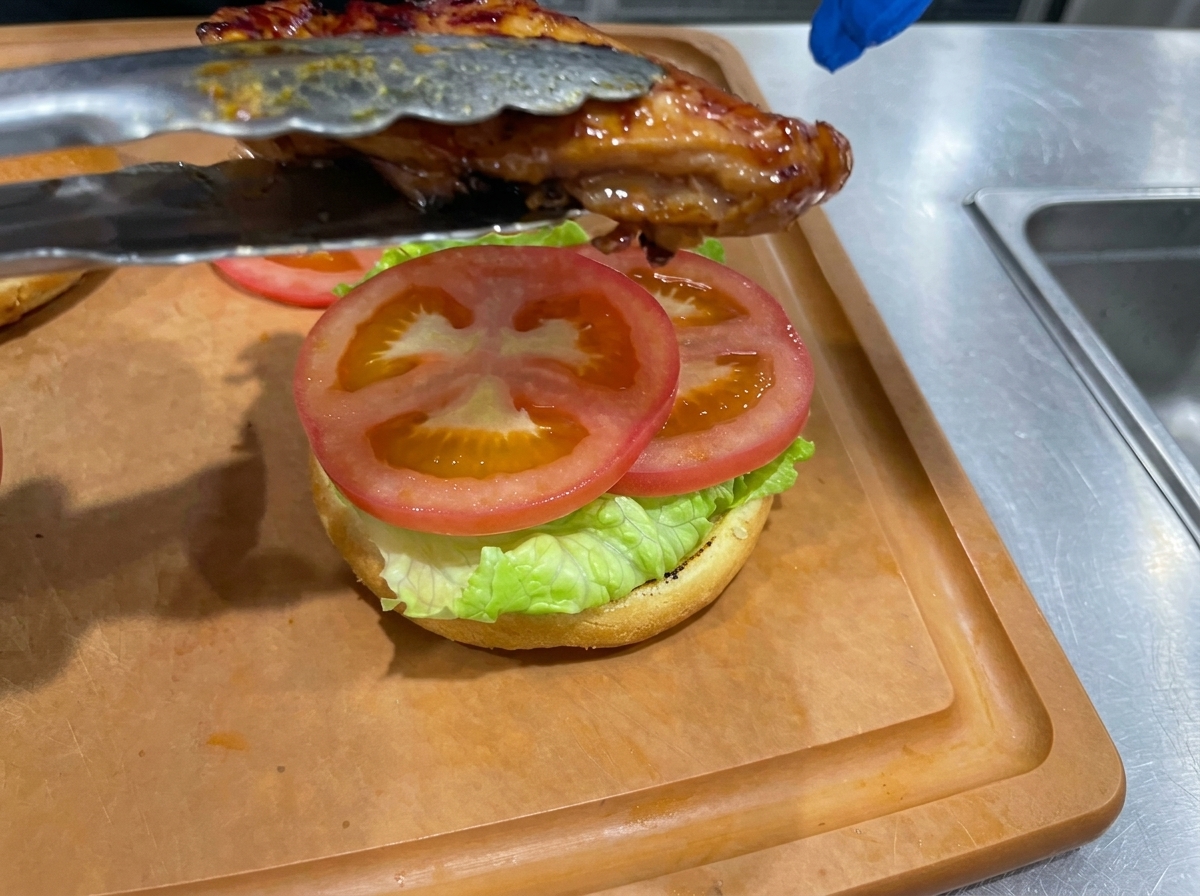 Usmażone udko z kurczaka w glazurze teriyaki układane szczypcami na bułce z sałatą i pomidorem.