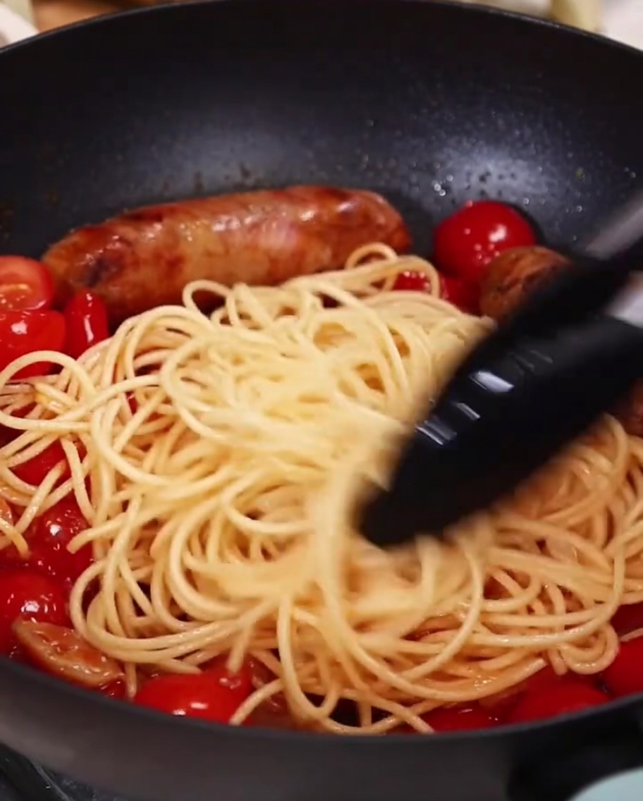 Spaghetti mieszane z pomidorami i kiełbasą na patelni za pomocą szczypiec.