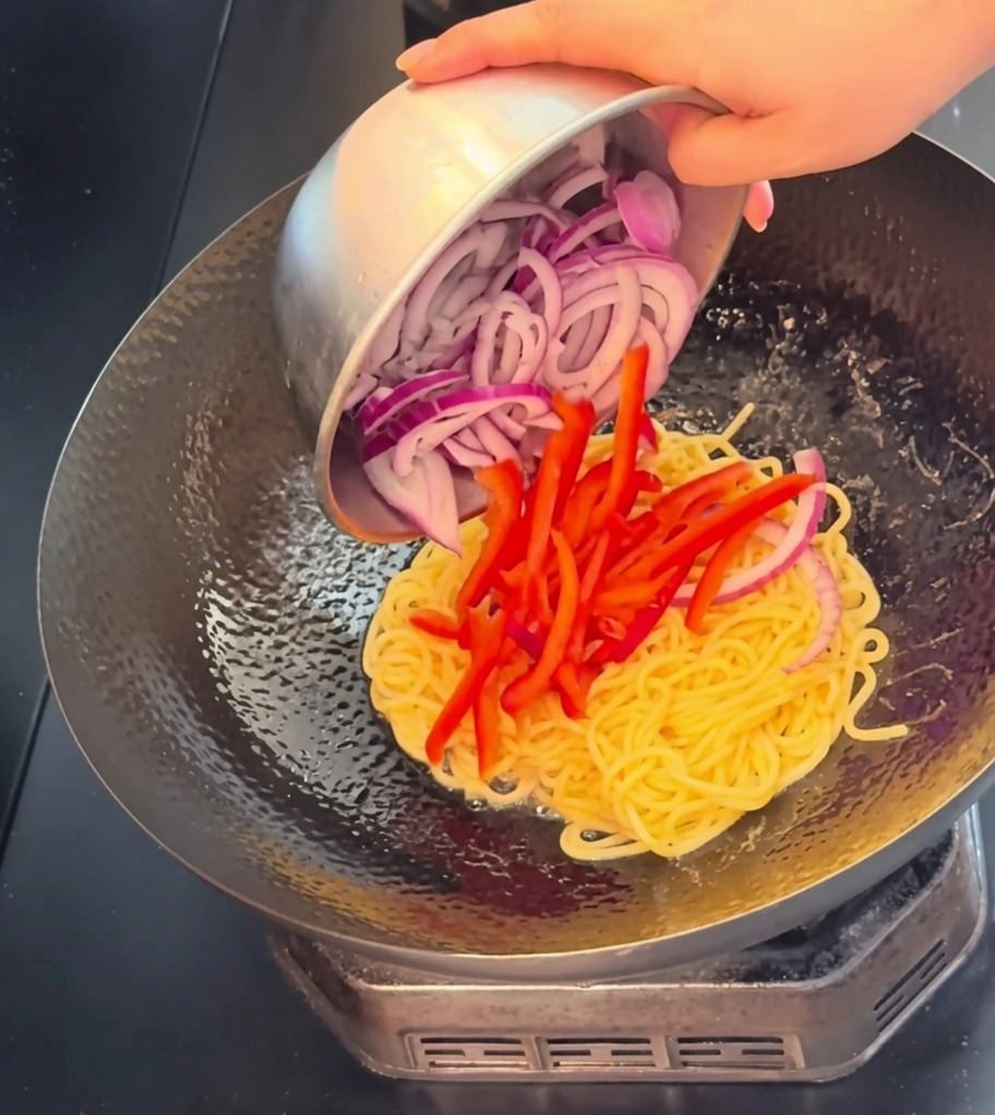 Zbliżenie na plastry wołowiny, paski czerwonej papryki i piórka czerwonej cebuli dodawane do woka ze spaghetti.