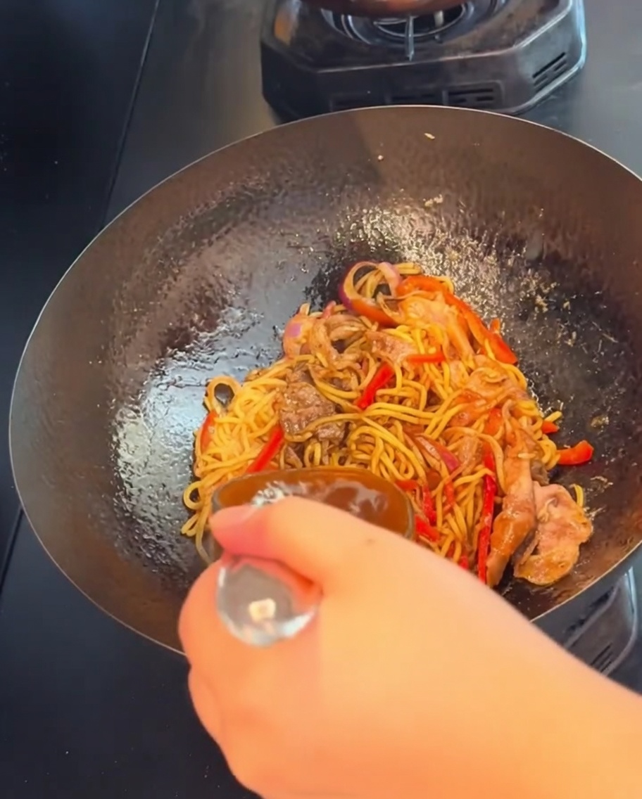 Ręka trzymająca łyżkę, energicznie mieszająca spaghetti z wołowiną w czarnym pieprzu podczas smażenia w woku.