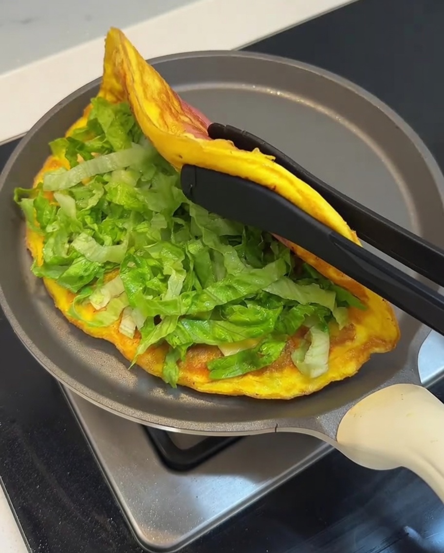 Omlet z placka cebulowego wypełniony sałatą, składany na pół na patelni za pomocą szczypiec.