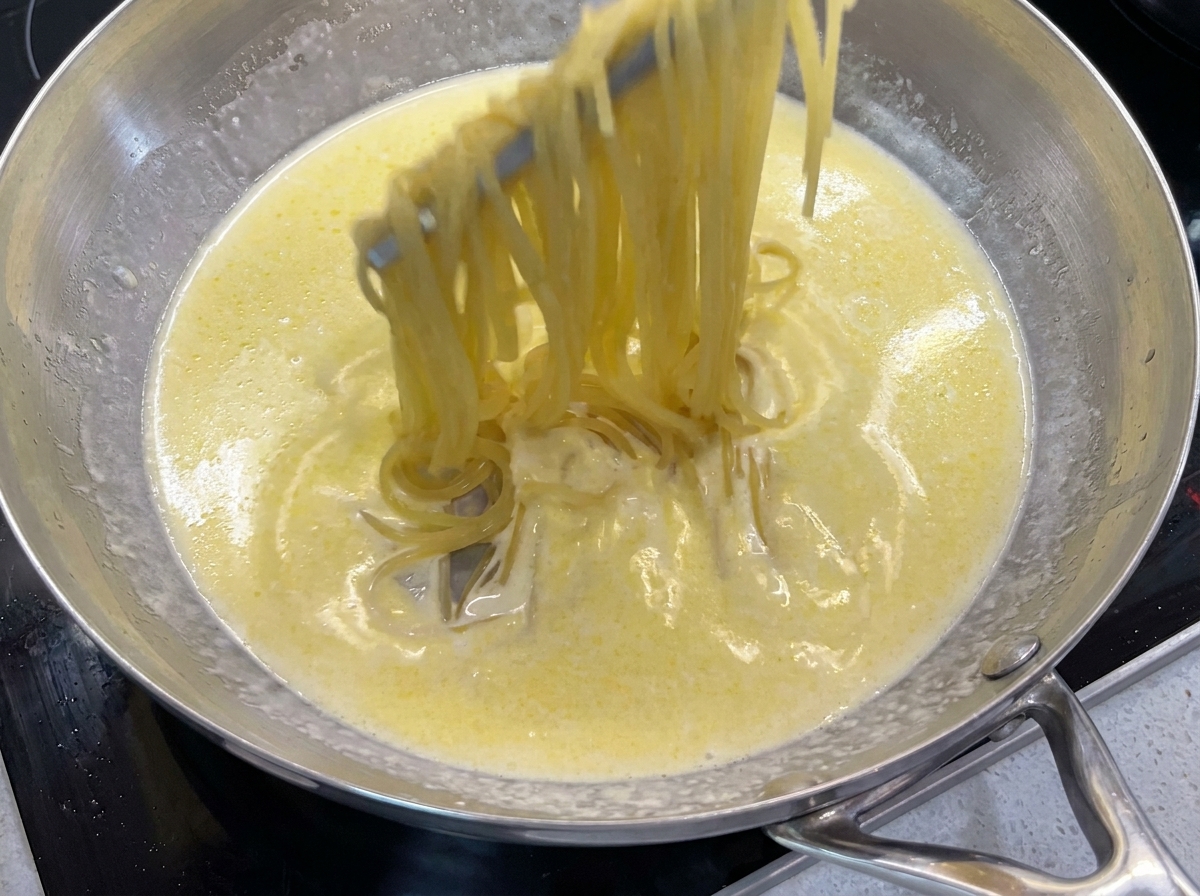 Metalowe szczypce mieszające ugotowane spaghetti w kremowym maślanym sosie na patelni.