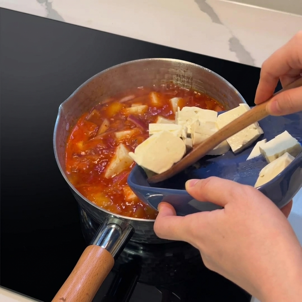 Pokrojone twarde tofu przekładane z niebieskiego talerza do wrzącego gulaszu.