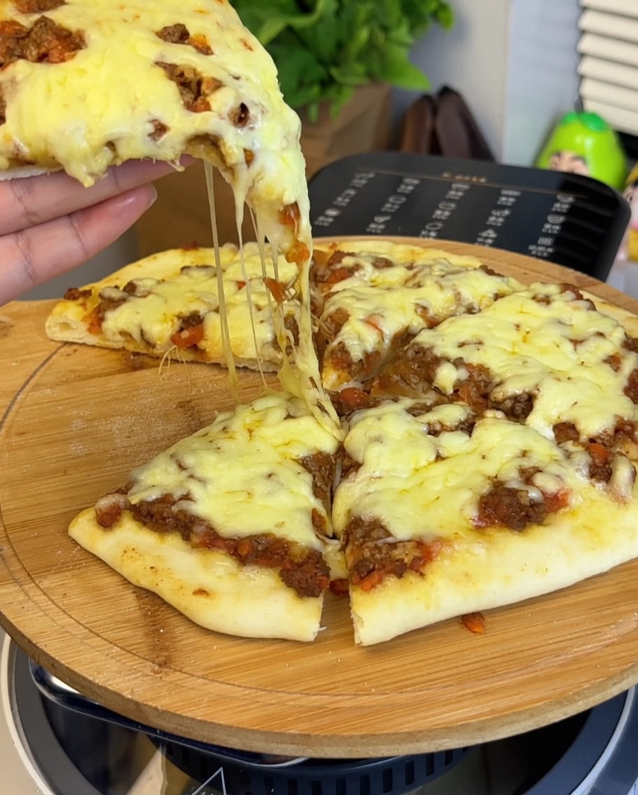 Zbliżenie na sos wołowo-pomidorowy rozprowadzany na spodzie do pizzy, z dłonią trzymającą kawałek pizzy w tle.
