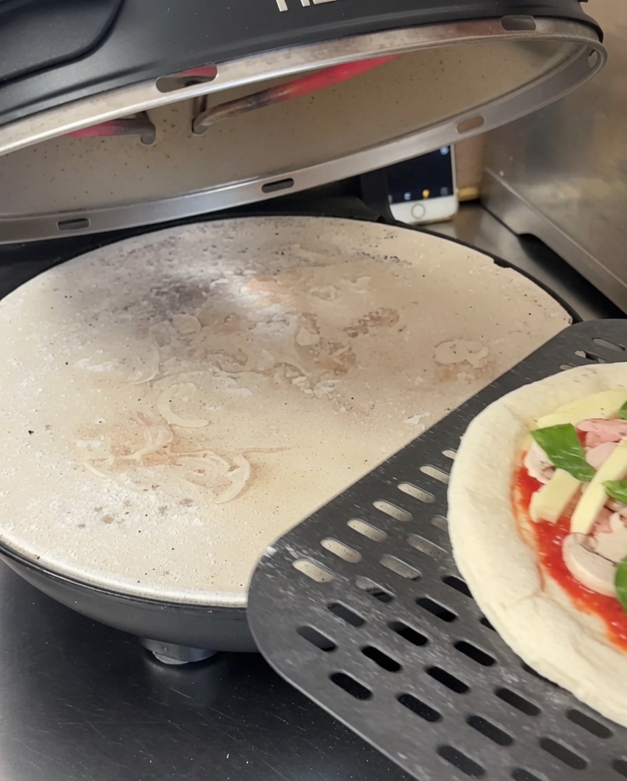 Spłaszczone ciasto na pizzę leżące na czarnym, perforowanym kamieniu w piecu do pizzy.
