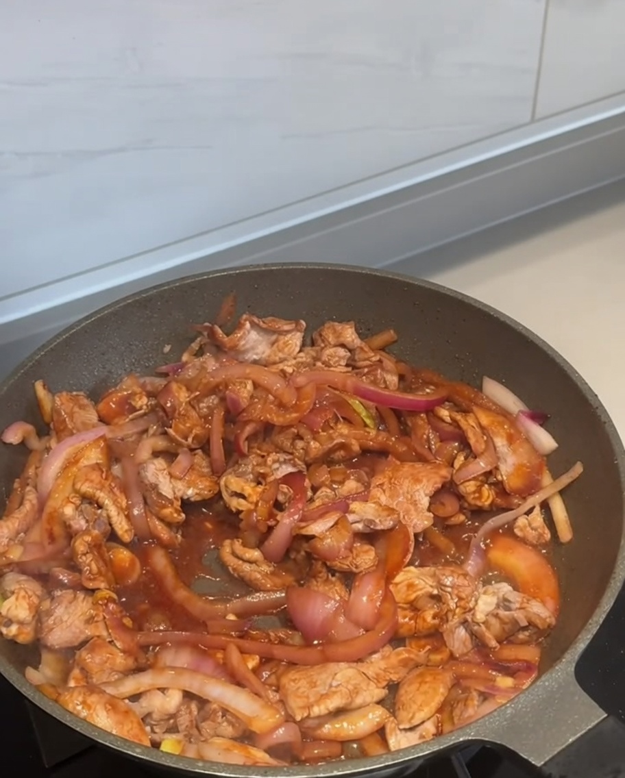 Płatki chili posypywane na smażącą się wieprzowinę z cebulą na patelni.