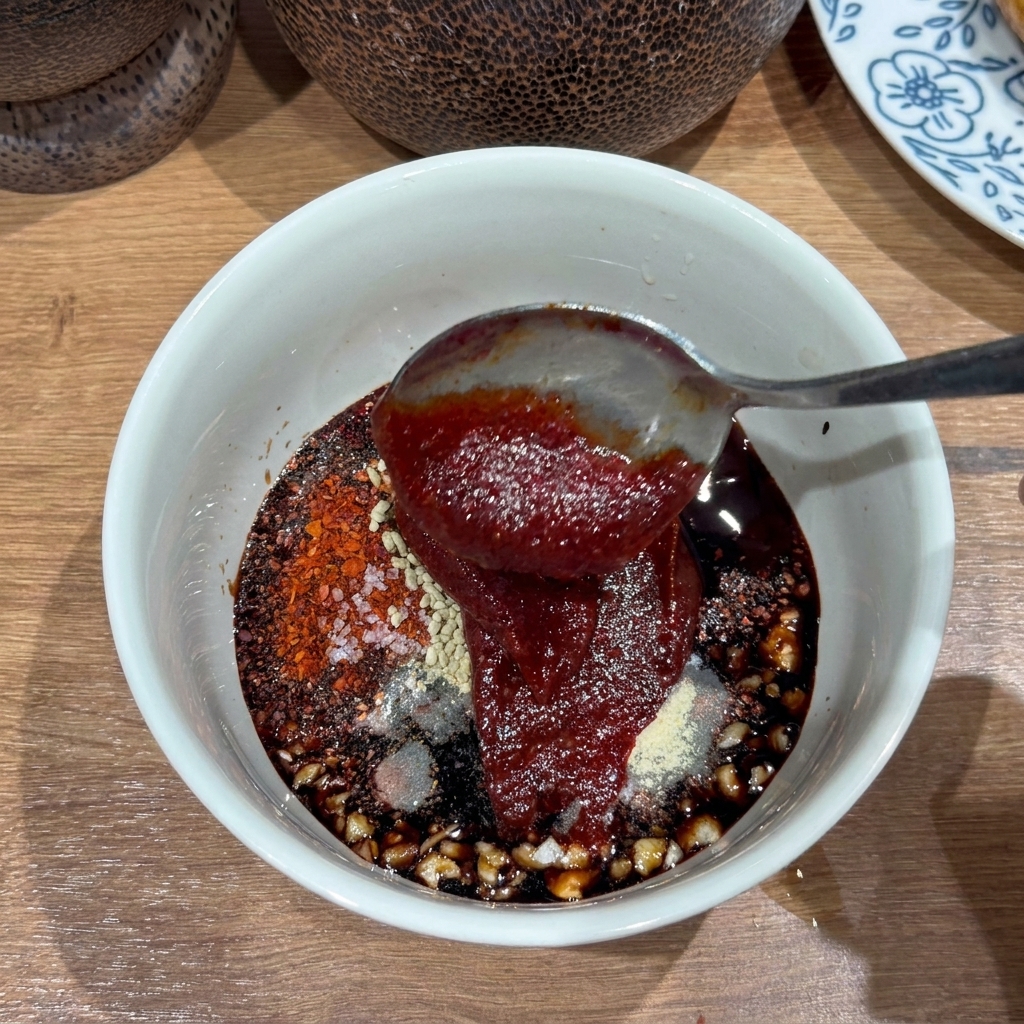 Zbliżenie na białą miseczkę z gęstym, ciemnoczerwonym sosem z widocznym czosnkiem i płatkami chili.