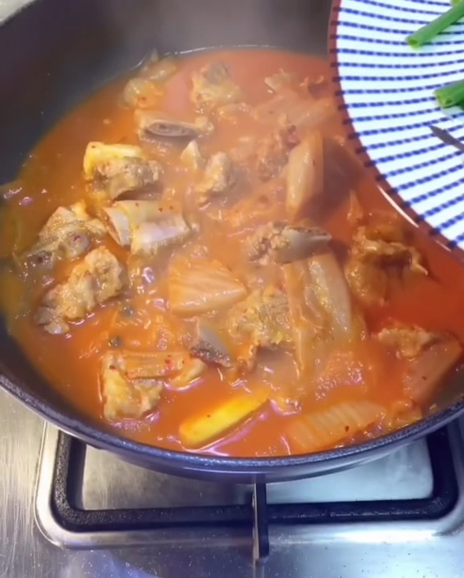 Zbliżenie na świeży szczypior, kolendrę i pokrojone zielone papryczki chili rozsypywane na bulgoczący, czerwony gulasz z kimchi i żeberkami.