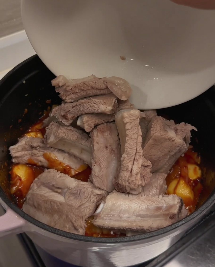 Zblanszowane wcześniej żeberka wieprzowe dodawane z białej miski do garnka z przyprawioną bazą kimchi.