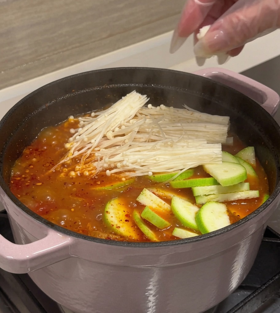 Plasterki cukinii, kostki tofu i grzyby enoki dodawane do garnka z czerwonym, bulgoczącym bulionem kimchi.