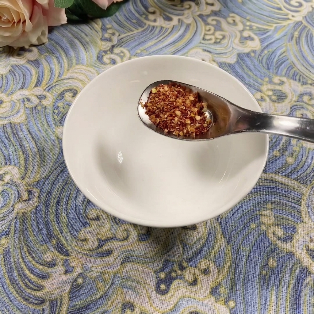 Łyżka grubo mielonego chili trzymana nad pustą białą miseczką na niebieskim wzorzystym tle.